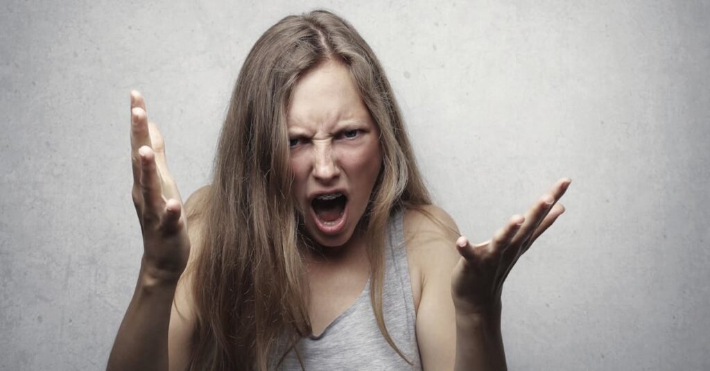 A young woman showing intense anger and frustration, yelling with hands raised against a gray background.